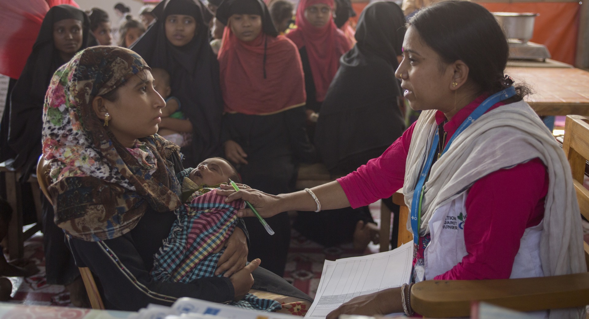 Health education officer, Shaki Rani Bose, talks to a young mother, Hosne Ara, with her baby, surrounded by Rohinga women as they wait patiently to be seen at an Action Against Hunger clinic for pregnant and breastfeeding mothers.